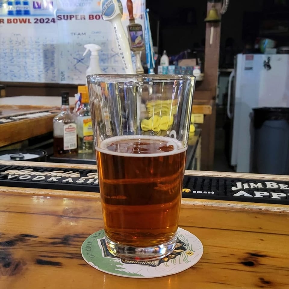 Pint of amber beer on a coaster at a rustic bar, with taps, bottles, and Super Bowl 2024 boards in the background.