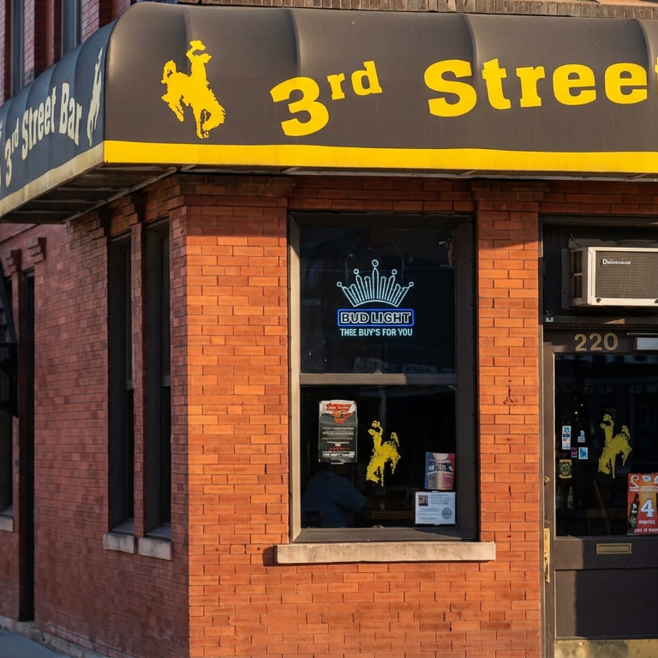 Brick-fronted 3rd Street bar entrance with black-and-yellow awning, cowboy logos, Bud Light window sign, and address 220.