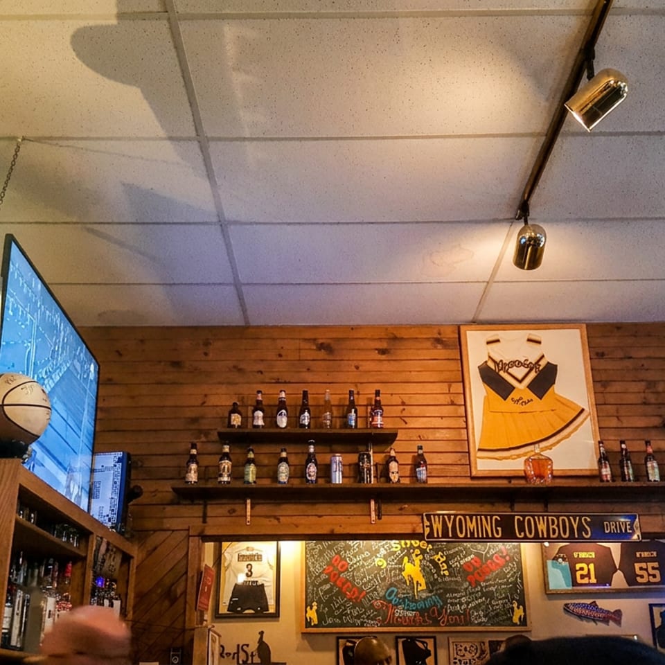 Rustic sports bar interior with wooden walls, liquor shelves, televisions, basketball, chalkboard, and Wyoming Cowboys memorabilia.